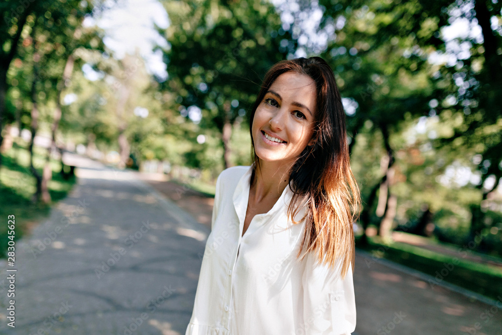 Caucasian woman with dark hair wearing white blouse walking un sunlight on background of summer park Caucasian woman with dark hair wearing white blouse walking un sunlight on background of summer park