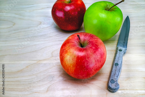 Three, red, green and yellow apples on a wooden table, knife with wooden handle, the background is a table made of light oak, close up