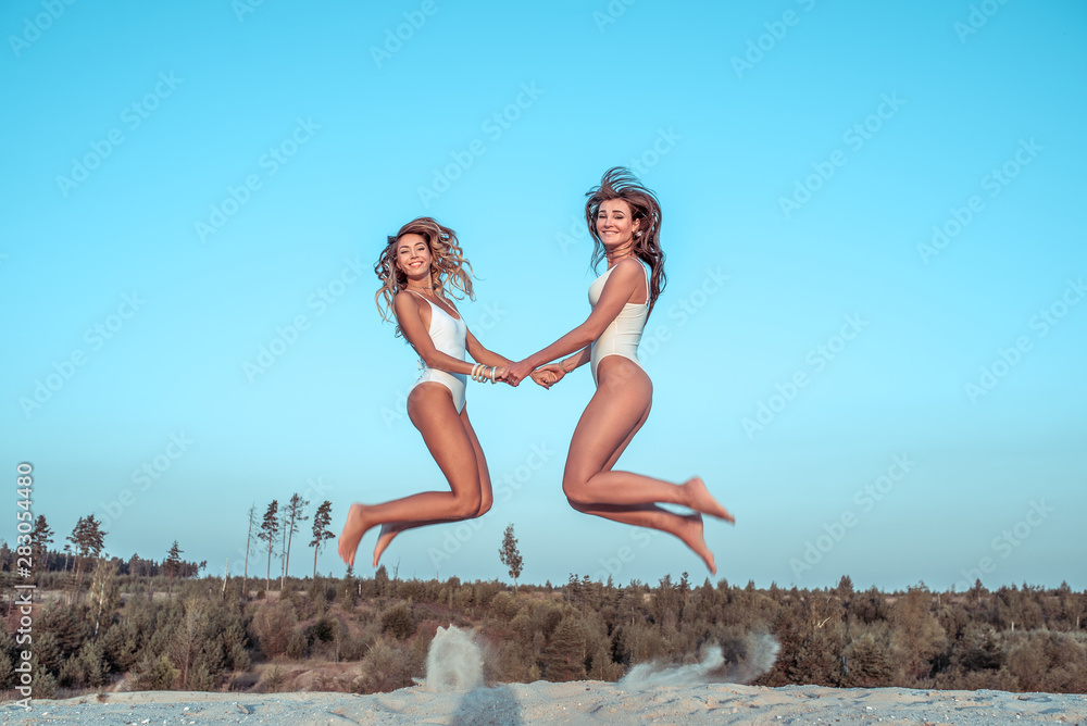 Two girlfriends in summer jump on sandy beach by sea, women relax in resort, tanned slim figures, white bodysuit. Stylish fashionable modern girls, happy, play, smile laugh, enjoy game and weekend. Two girlfriends in summer jump on sandy beach by sea, women relax in resort, tanned slim figures, white bodysuit. Stylish fashionable modern girls, happy, play, smile laugh, enjoy game and weekend.