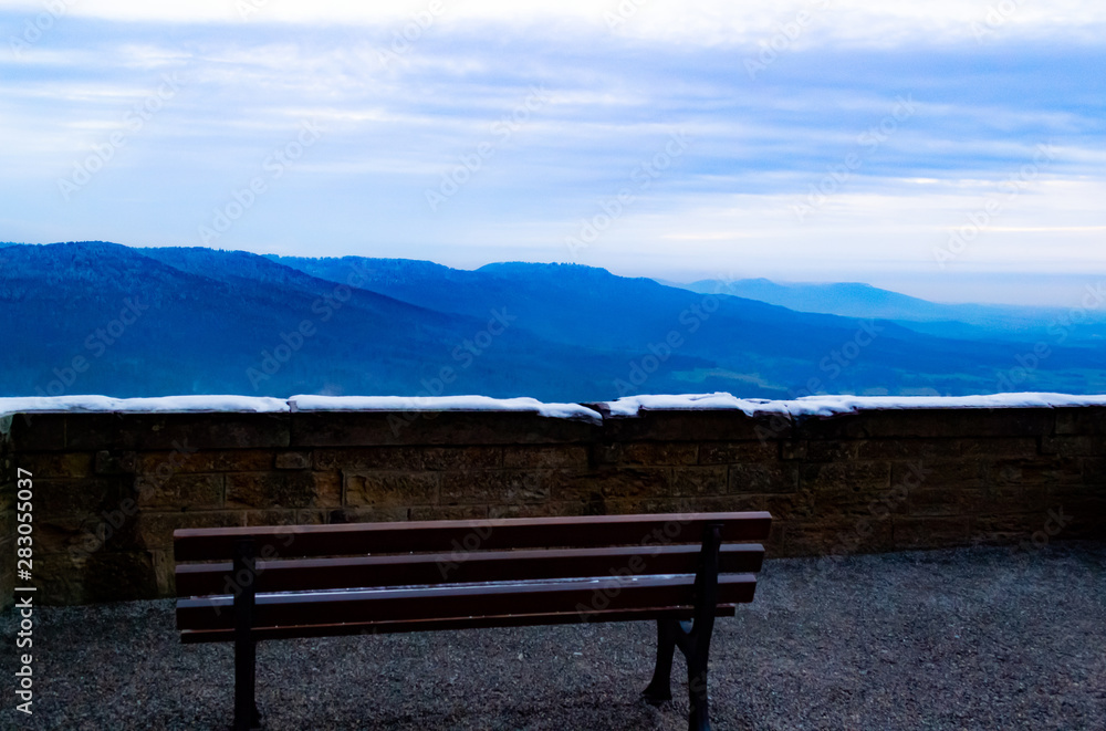 High in the mountains, in the courtyard of Hohenzollern castle is a lonely bench overlooking the mountains, blue sky and fluffy white clouds. Daytime.