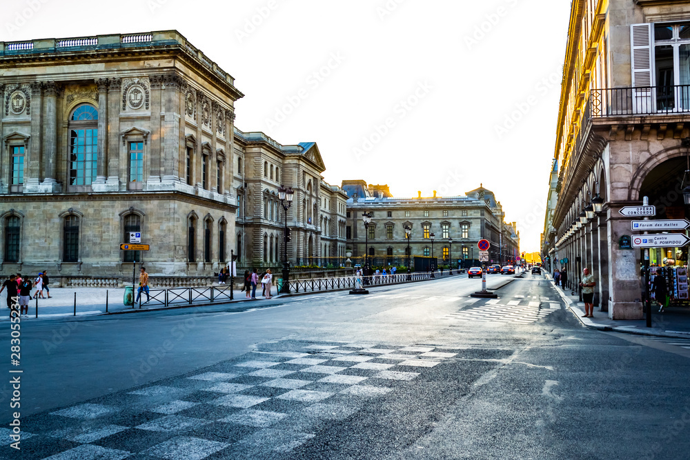 Paris Louvre Museum. Main entrance. The facade of a beautiful historic building. Asphalt road on which the car ride. Pedestrian crossing, people walk along the sidewalk. Street lights and pointers