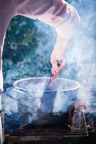 Female hand stir food in a cauldron on a background of smoke from the fire © SerPhoto