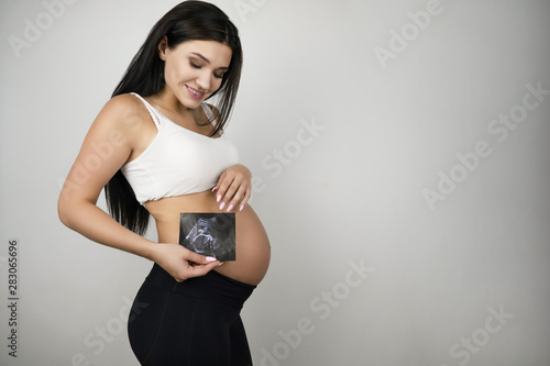 beautiful pregnant brunette woman holding ultrasound picture of her baby on isolated white background