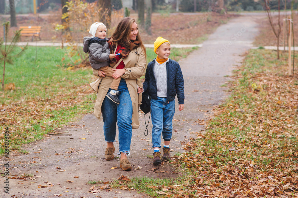 Fototapeta premium Young mother taking child to school, and baby son to kindegarden. Autumn time change. Parent, baby son, son schoolboy. Happy family on a walk in autumn park