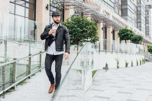 A bearded, serious, stylish man walking  on the streets of the city near modern office centre and drinks coffee. © Тарас Нагирняк
