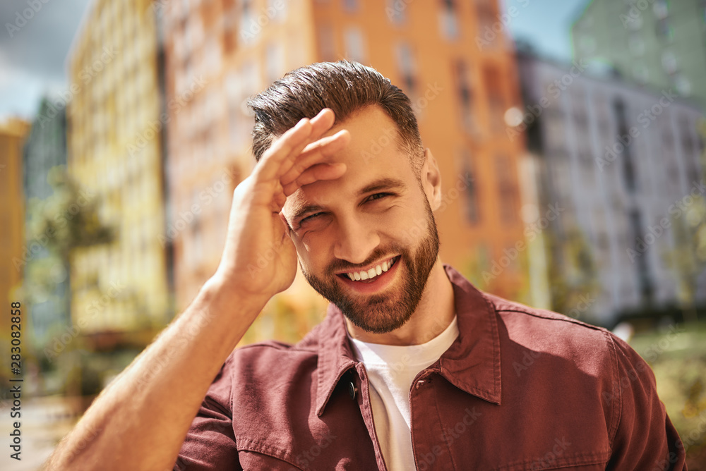 Sunny day Portrait of happy attractive man with stubble smiling and looking at camera while standing on the street Sunny day Portrait of happy attractive man with stubble smiling and looking at camera while standing on the street