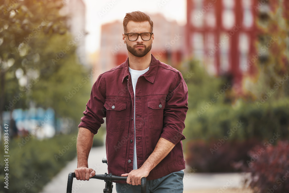 Perfect day for cycling. Handsome man with stubble in casual clothes and eyeglasses pulling his bicycle and looking at camera while walking outdoors