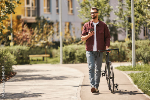 My way. Handsome man with stubble in casual clothes and eyeglasses pulling his bicycle and holding smartphone while walking outdoors