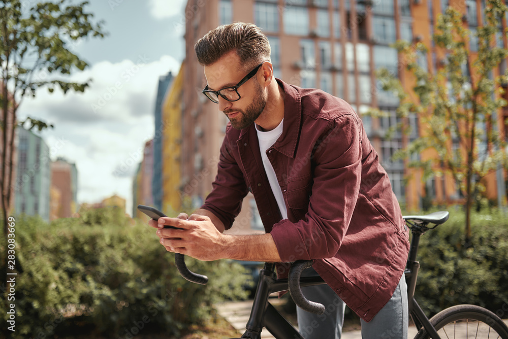 Surfing the net. Side view of handsome man with stubble in casual clothes and eyeglasses leaning at his bicycle and looking at mobile phone while standing outdoors Surfing the net. Side view of handsome man with stubble in casual clothes and eyeglasses leaning at his bicycle and looking at mobile phone while standing outdoors