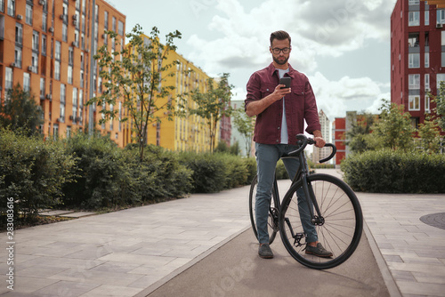 Fototapeta Naklejka Na Ścianę i Meble -  Chatting with friend. Full length of handsome man with stubble in casual clothes and eyeglasses holding mobile phone while walking with his bicycle outdoors