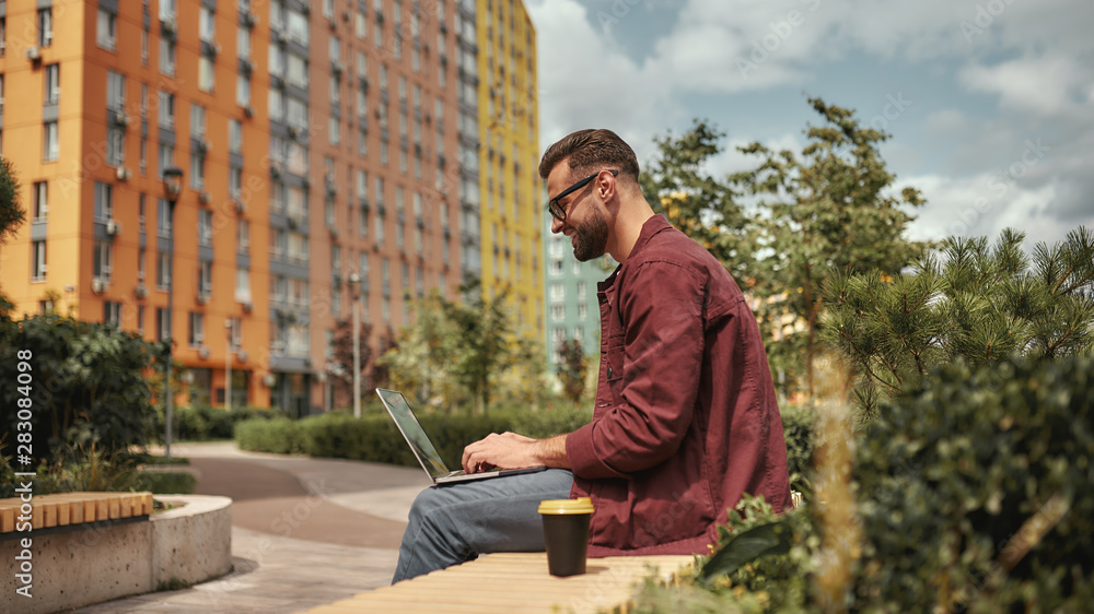 Happy and young. Side view of handsome man with stubble in casual clothes and eyeglasses working on laptop while sitting on the bench outdoors Happy and young. Side view of handsome man with stubble in casual clothes and eyeglasses working on laptop while sitting on the bench outdoors