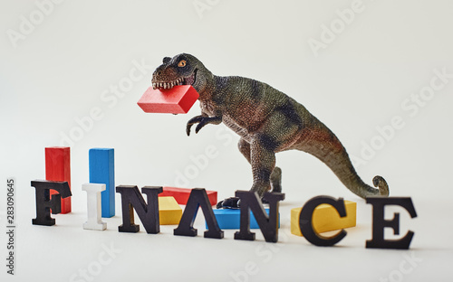 Finance word created with alphabet letters. Dinosaur toy standing with a red block in a mouth near multi-colored wooden blocks in the background