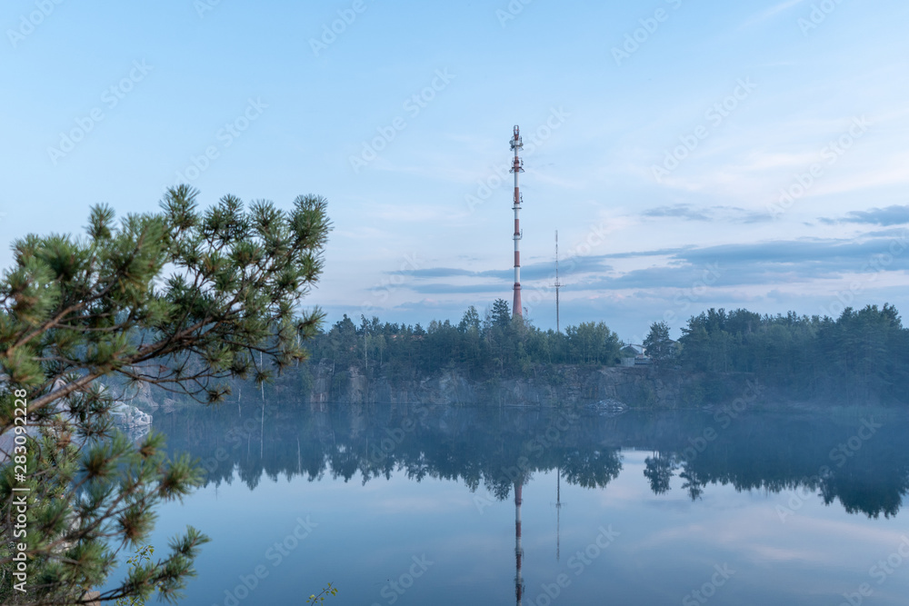 Picturesque landscape. Dramatic scenic view of the granite quarry filled with blue water, and the opposite shore overgrown with trees after sunset at dusk in the summer. High tower on the bank. 