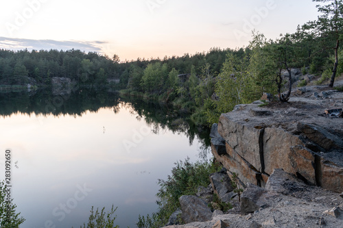 Dramatic scenic panorama of the granite quarry filled with water, and the opposite rocky shore overgrown with after sunset at dusk in the summer. 