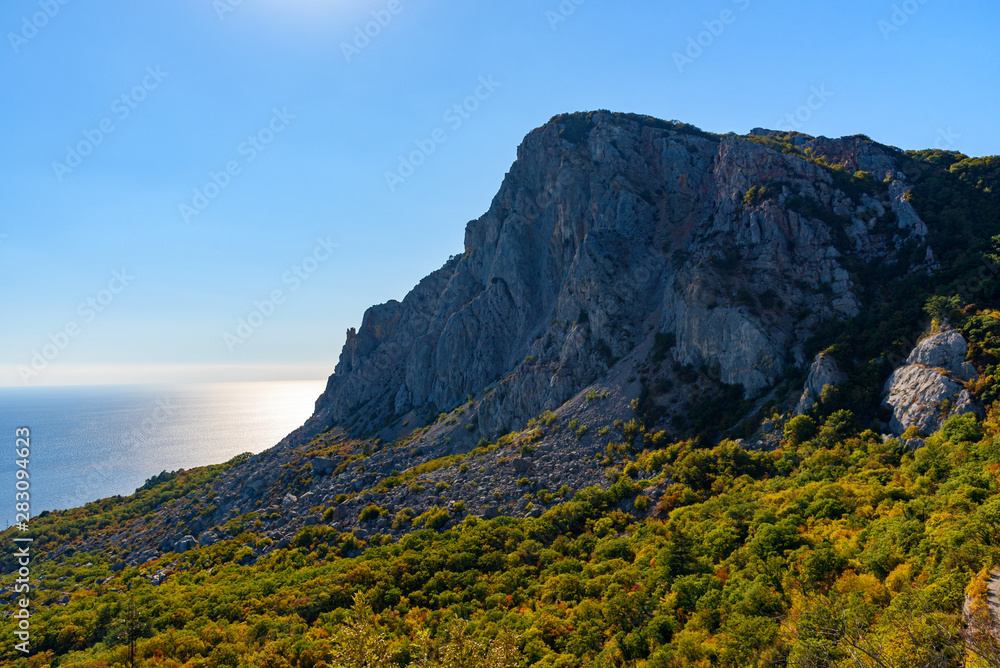 view of the stony slope of the mountain at the foot of which trees grow, on the shore of the Black Sea on a bright sunny cloudless day.