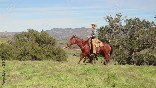 Sitting tall on his horse, the cowboy spurs his horse to move through the frame as the camera dolly's towards a barn in the distance photo