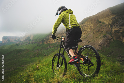 Aged male athlete in helmet and mask rides a mountain bike on a grassy slope against the background of plateau rocks and low clouds on an overcast day. Downhill mountain bike concept