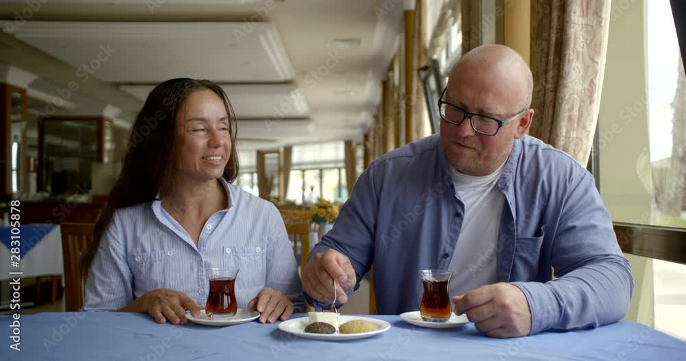 A bald man in glasses is having Breakfast with his wife in the hotel restaurant to have fun and tries to feed dessert with tea A bald man in glasses is having Breakfast with his wife in the hotel restaurant to have fun and tries to feed dessert with tea