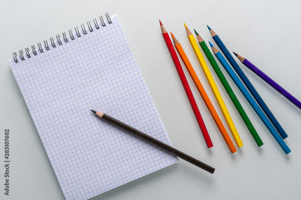 Clean notepad in a cage with a pencil lying on it and rainbow color pencils on a white background top view copy space Clean notepad in a cage with a pencil lying on it and rainbow color pencils on a white background top view copy space