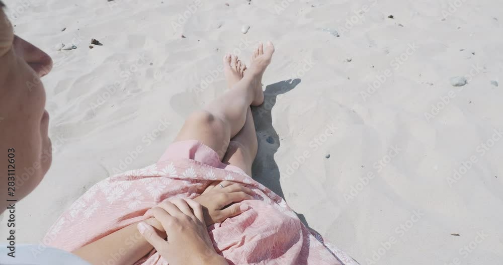 Woman resting on sandy beach. Wind sways light fabric of pink skirt. Woman holds the clothes with her tanned hands. Shooting from behind shoulder. Woman resting on sandy beach. Wind sways light fabric of pink skirt. Woman holds the clothes with her tanned hands. Shooting from behind shoulder.