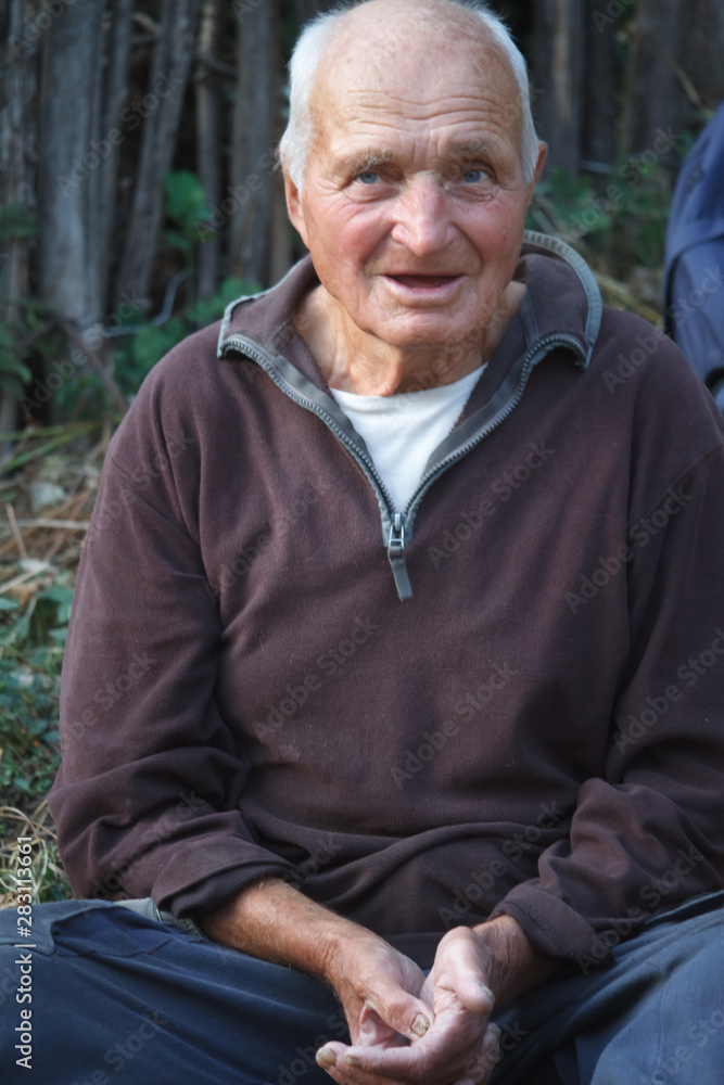 Close-up portrait of a very old man with arms crossed, sits on the ground on a blurred background of wood fence, selective focus Close-up portrait of a very old man with arms crossed, sits on the ground on a blurred background of wood fence, selective focus