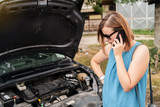 Young woman tourist driver standing by the road by the car with the hood open dead engine broken automobile malfunction repair in a summer day making a mobile phone call for help