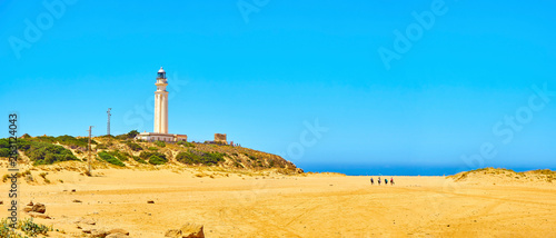 Tourists walking in direction to Faro de Trafalgar Beach, a broad beach of The Cabo de Trafalgar Cape Natural Park with the famous Lighthouse in the background. Barbate, Los Caños de Meca, Cadiz. Anda photo