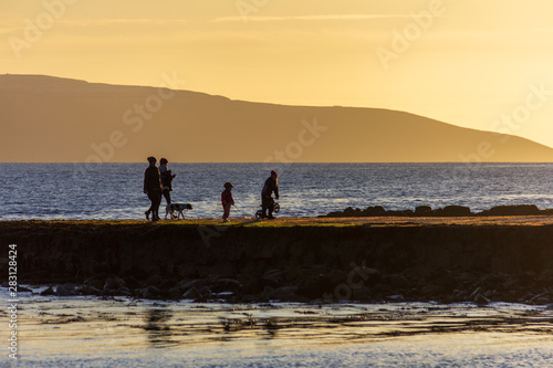 A family walking on a Salthill, Galway, Ireland jetty at sunset appear in silhouette against a yellow sky at sunset