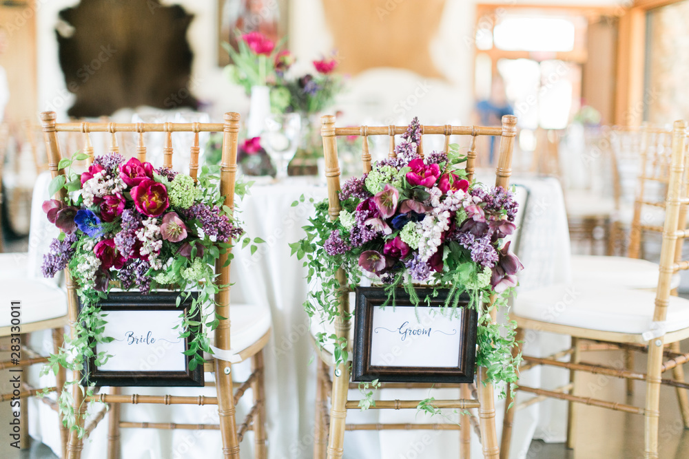 Bride and groom chair decorated with flowers and sign, back of chairs at wedding reception