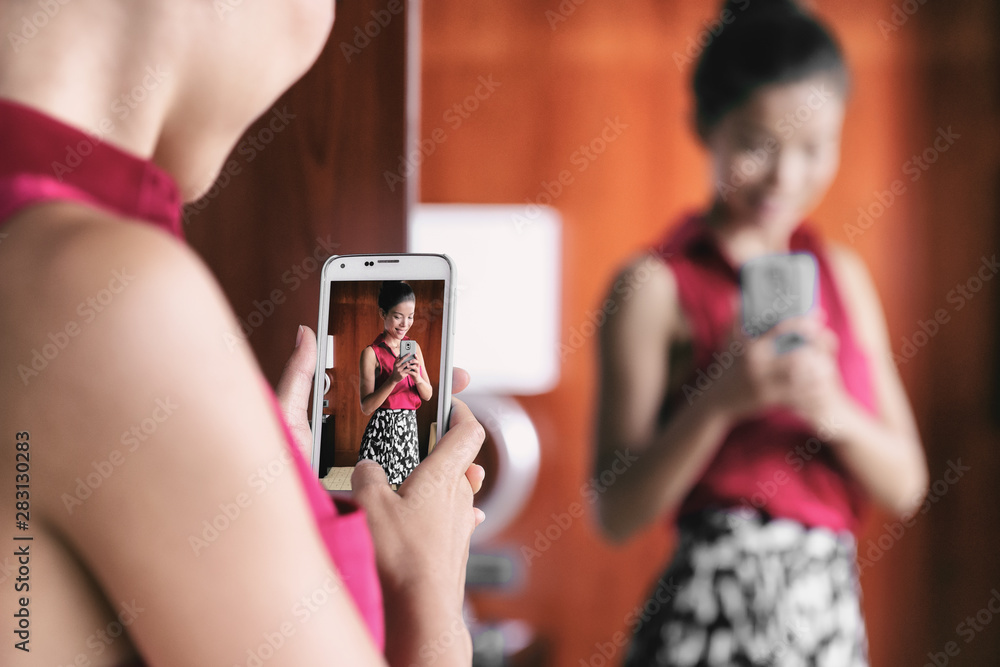 Girl turn attention at camera, distracted from smartphone smiling friendly  and happily holding mobile phone as, taking selfie in mirror, showing  friends new outfit, standing in dressing room of mall. Photos