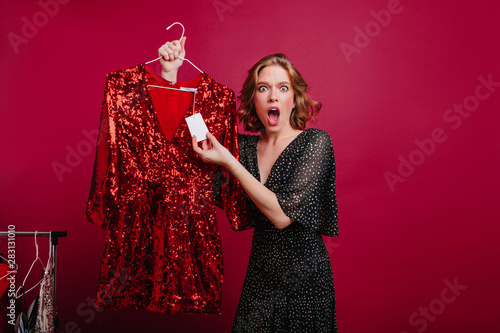 Young caucasian woman with shocked face expression looking at price on sparkle dress. Unhappy girl with short haircut holding hanger with expensive attire in store. photo