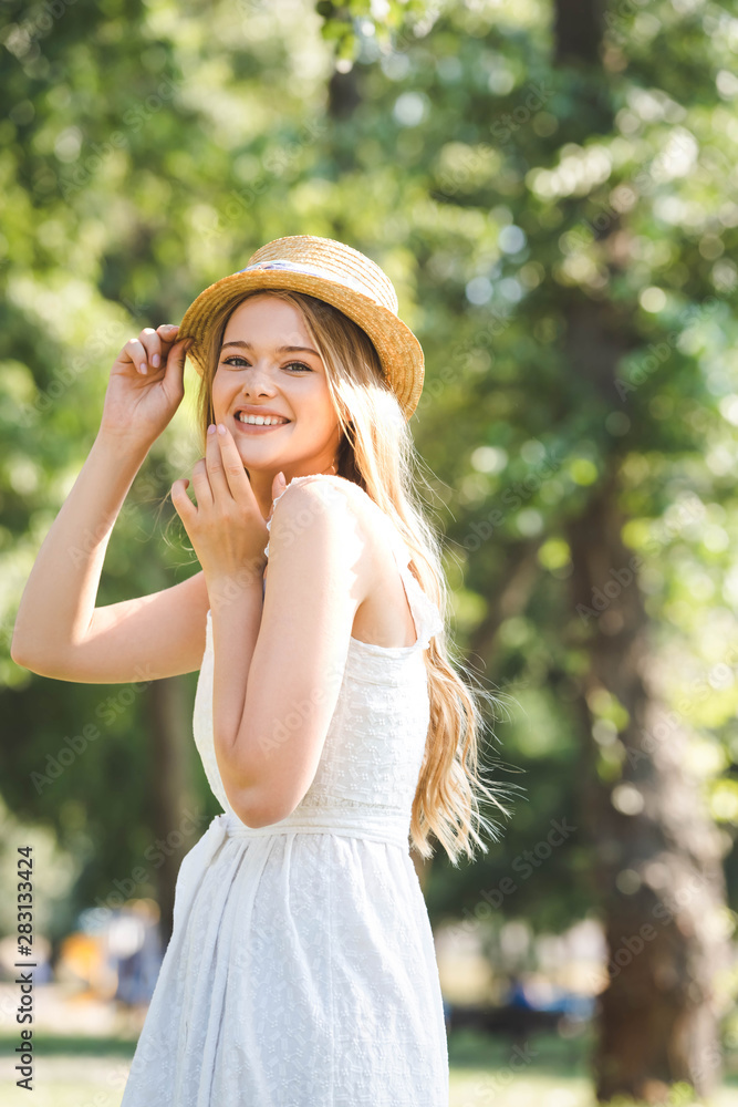 selective focus of beautiful young woman in white dress and straw hat smiling and looking at camera