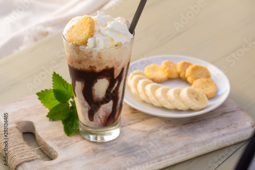 Tasty milkshake drink in glass mug with plastic tube and slice of banana on chopping board on wood table