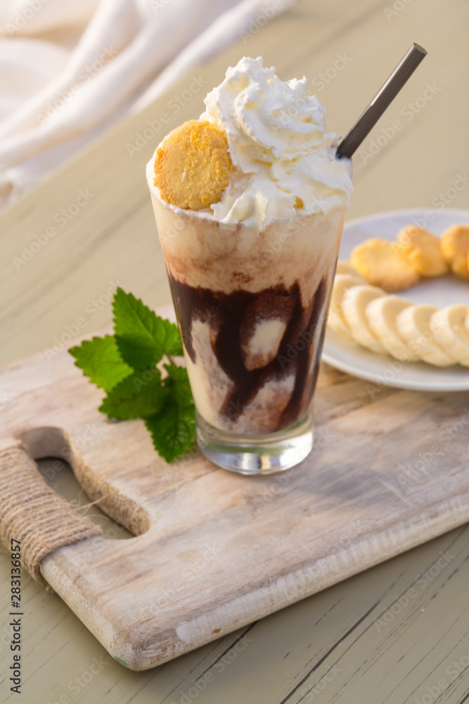 Tasty milkshake drink in glass mug with plastic tube and slice of banana on chopping board on wood table Tasty milkshake drink in glass mug with plastic tube and slice of banana on chopping board on wood table