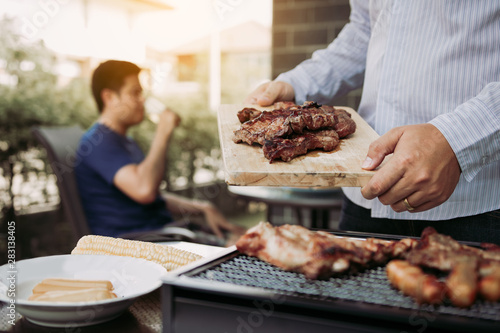 Asian men are preparing pork that is placed on a wooden cutting board and holding it to friends who are celebrating in the back.