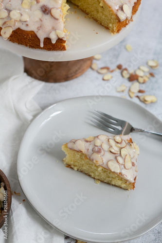 slice of almond olive oil cake with brown butter icing and sliced almonds, dripping icing on cake, marble and wood cake stand, styled bakery
