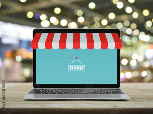 Modern laptop computer with online shopping store graphic and open sign on wooden table over blur light and shadow of shopping mall, Business internet shop online concept