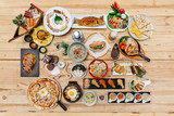 Flatlay of International Foods on wooden table: Sushi, Sashimi, Nachos, Spaghetti, Naan, Sandwich, Noodle, Steak, Ramen. Yakitori, Pizza, Bibimbap, Fried Chicken Wings, Wonton Soup and Brownie Frappe.