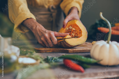 Female hands cutting pumkin on dark wooden kitchen table with vegetables cooking ingredients.