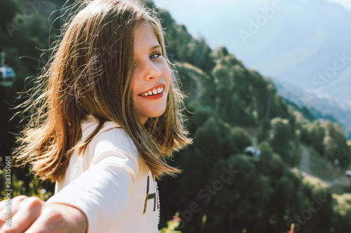 Smiling tween girl reaches out to camera follow me on background of beautiful mountains, family travel concept