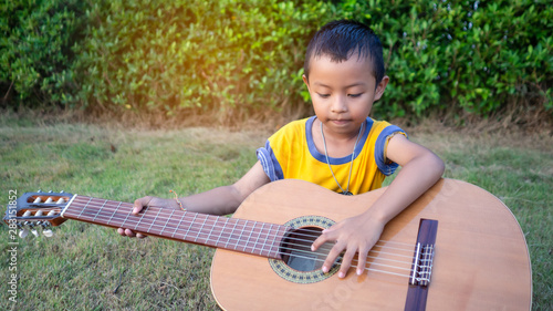  An Asian boy is playing an acoustic guitar in a garden with green trees.