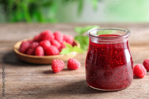 Glass jar of sweet jam with ripe raspberries on wooden table. Space for text