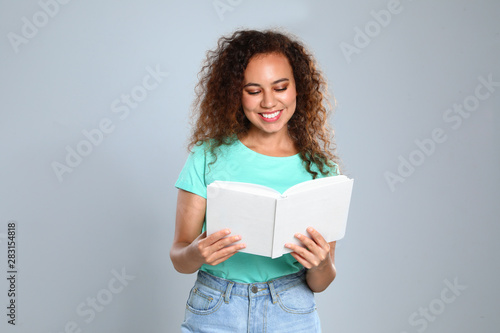 Beautiful African-American young woman reading book on light background