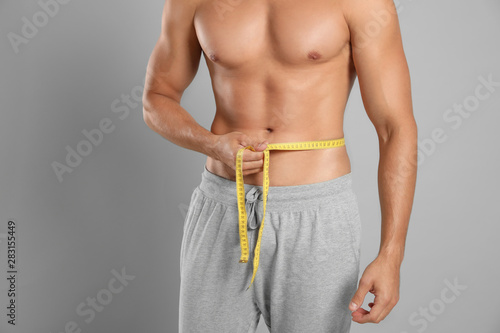 Young man with slim body using measuring tape on grey background, closeup view