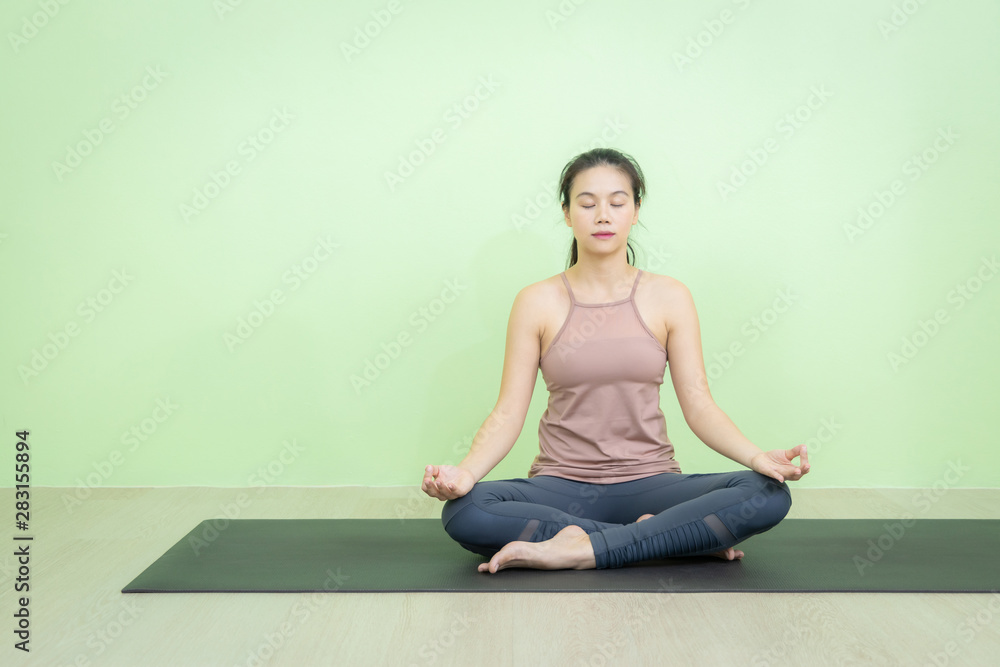 Beautiful smiling Asian woman sitting on black mat in lotus posture yoga, beginning relaxation for meditation and exercise in the room with green background wall. Beautiful smiling Asian woman sitting on black mat in lotus posture yoga, beginning relaxation for meditation and exercise in the room with green background wall.
