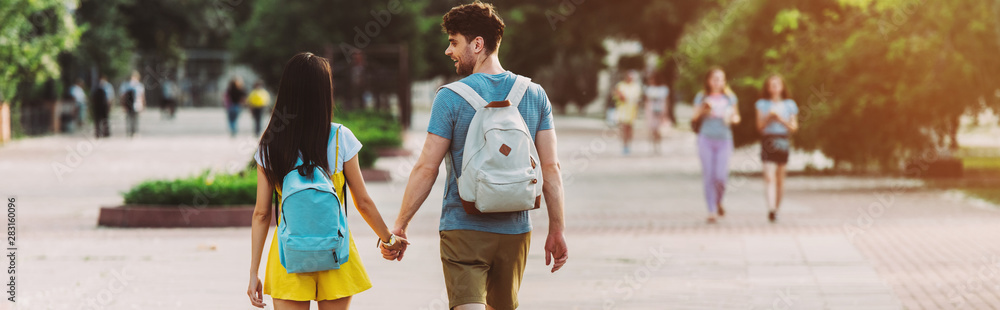 panoramic shot of man and woman with backpacks walking and holding hands - obrazy, fototapety, plakaty 