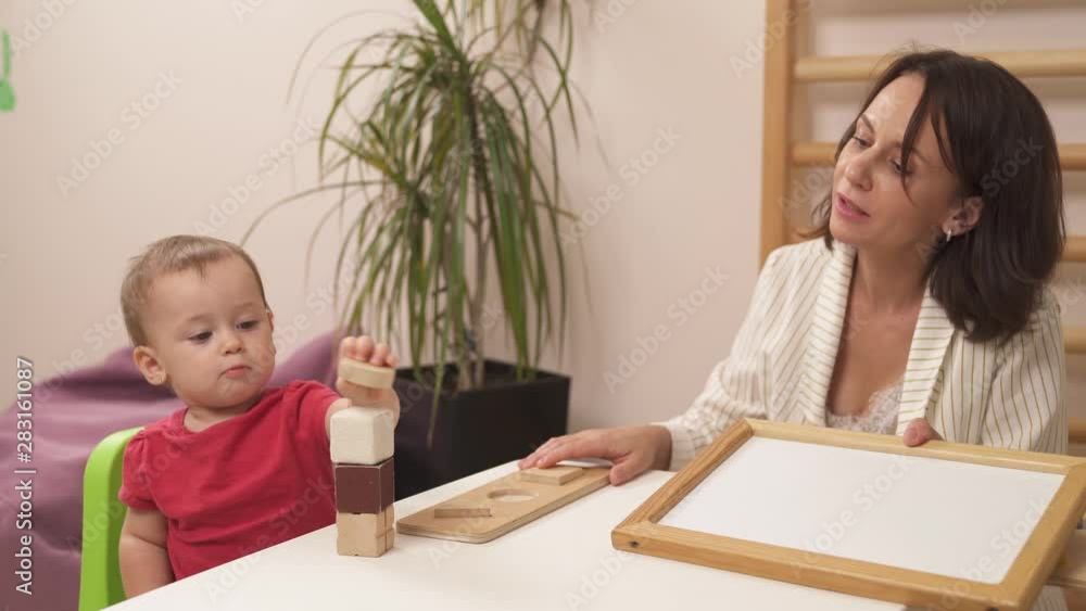 Close-up of a young therapist playing with her toddler patient by building a tower from wooden blocks. Development of kids fine motor skills Close-up of a young therapist playing with her toddler patient by building a tower from wooden blocks. Development of kids fine motor skills