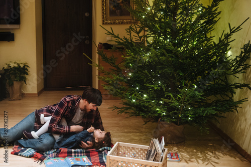 Family portrait of father and daughter in Christmas clothes having fun on the floor and christmas tree near them