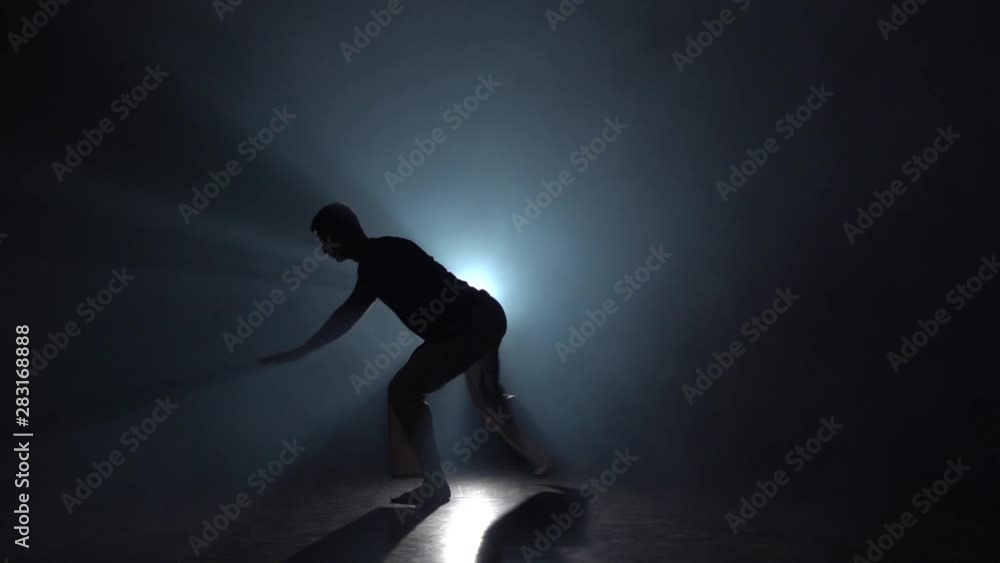 Silhouette male in darkness against light spotlight practicing capoeira . Slow motion. Silhouette male in darkness against light spotlight practicing capoeira . Slow motion.