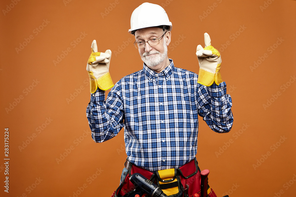Happy mature retired male construction worker wearing yellow rubber gloves, waist bag and white hardhat looking at camera with joyful broad smile, ready for work, pointing both index fingers upwards Happy mature retired male construction worker wearing yellow rubber gloves, waist bag and white hardhat looking at camera with joyful broad smile, ready for work, pointing both index fingers upwards